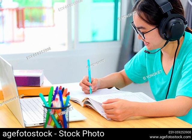 Asian woman student teenage girl with glasses headphones sitting happy smile looking writing notes at book laptop computer on table learning online study...