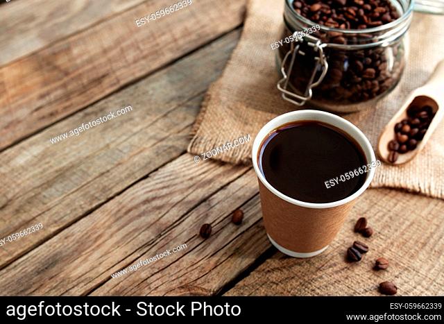 Concept of breakfast in cafe with paper cup of black coffee espresso, dry roasted coffee beans in the glass jar and spoon at old wooden table