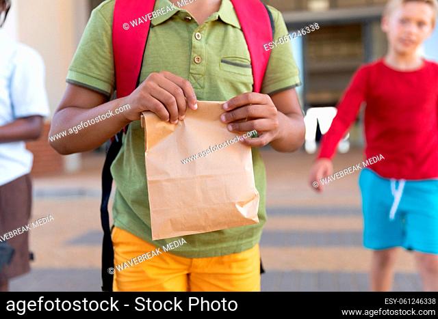 Midsection of biracial elementary schoolboy with packed lunch standing in school campus