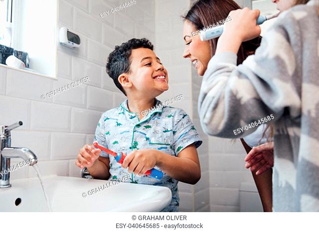 Children are brushing their teeth in the bathroom at home. The mother is checking the little boy's mouth to make sure he has brushed properly