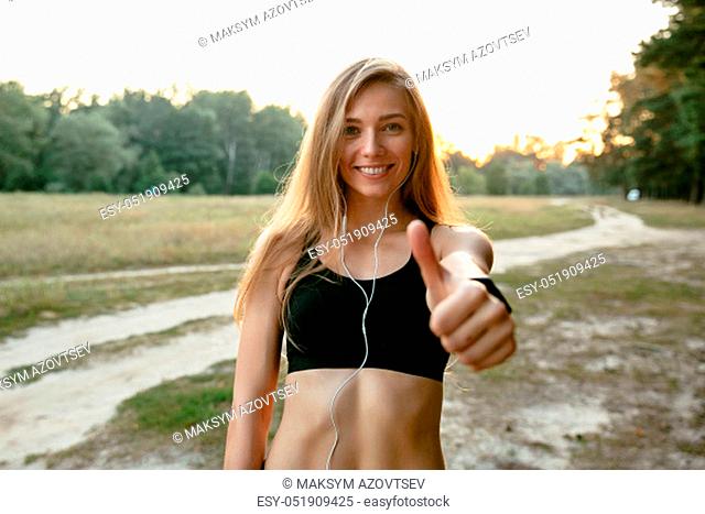 Beautiful smiling girl in earphones, showing a thumb up and looking at camera, wearing sportswear. Outdoors