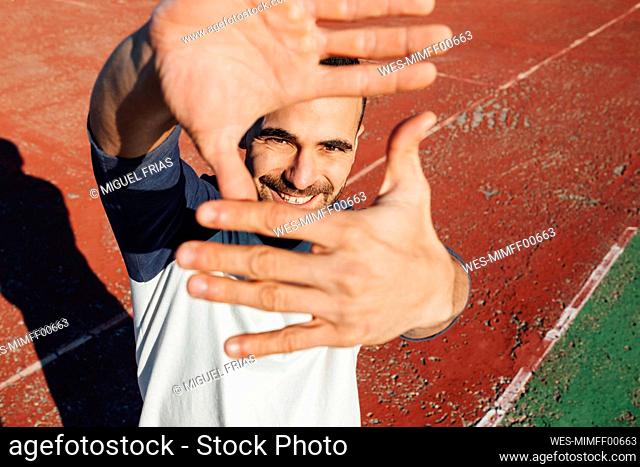 Smiling man looking through finger frame on sports court during sunny day