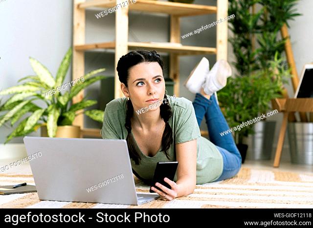 Contemplating woman holding smart phone by laptop while lying at home