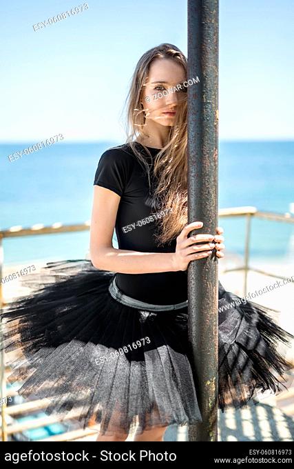 Attractive ballerina holds a tube on the concrete pier on the clear sky and sea background. She wears a black tutu with a leotard