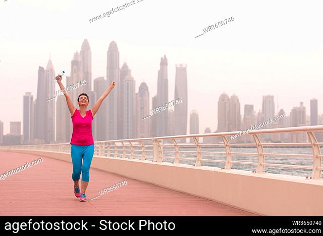 young woman celebrating a successful training run on the promenade by the sea with a bottle of water and her hands raised in the air with a big city in the...