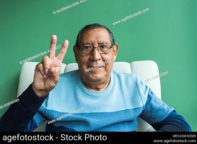 Senior man gesturing peace sign in front of green wall