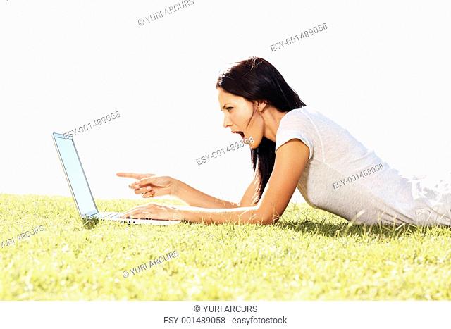 Side view of surprised young woman pointing at laptop screen while lying down in grass