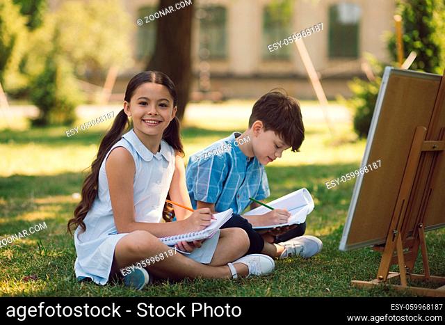 Happy Children Writing Tasks Outside While Sitting On Grass On Sunny Day. Education In Nature Concept