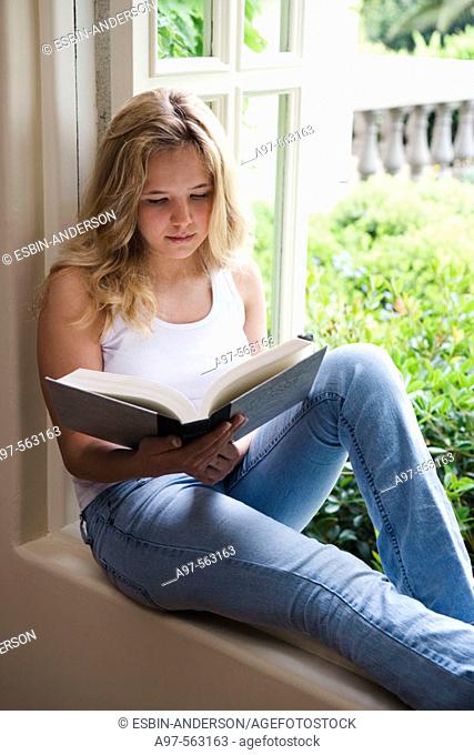 Blonde barefoot teen girl sits by an open window reading a book