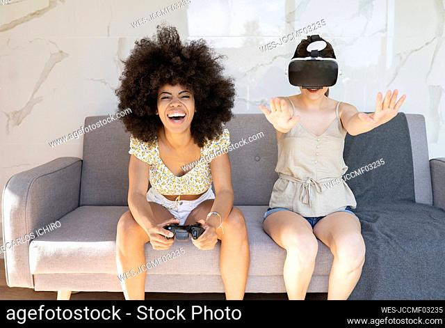 Happy woman holding joystick sitting on sofa with female friend wearing virtual reality headset at home