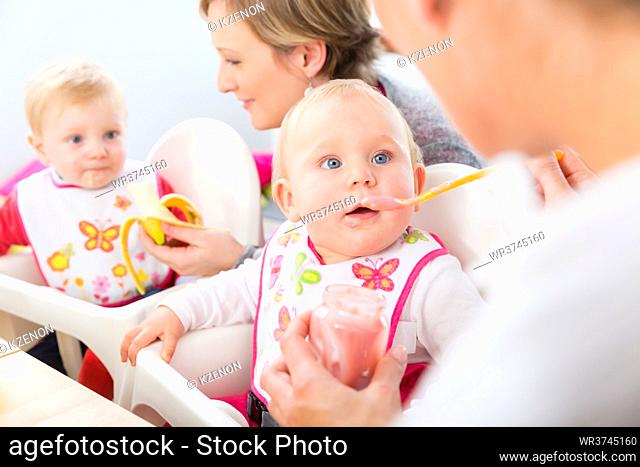 Portrait of a cute and healthy baby girl with blue eyes looking at her mother, while eating nutritious fruit puree next to another baby at home