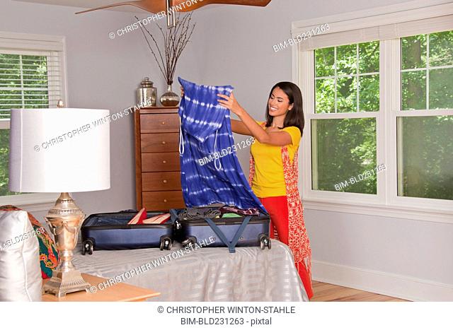 Smiling Hispanic woman examining dress over suitcase in bedroom