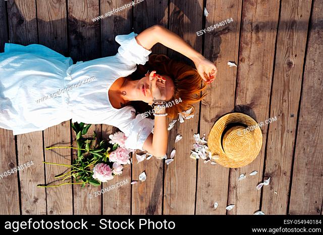 Attractive young hipster woman in white dress and straw hat lies on the wooden floor
