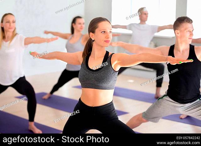 Attractive yogi woman and a group of young sporty people practicing yoga lesson, doing Warrior Two exercise, Virabhadrasana II pose, working out indoor