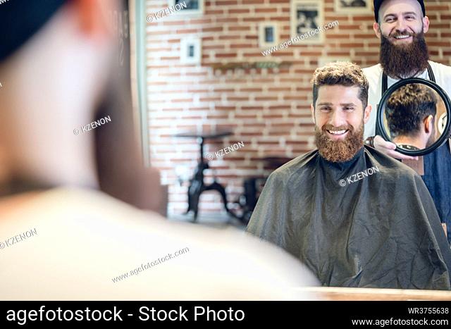 Over the shoulder view of a handsome young man smiling, while looking at his new trendy haircut in the mirror held by his experienced barber in a cool hair...