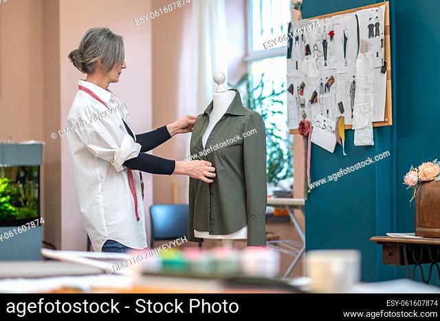 Creative process. Focused gray-haired serious woman standing sideways to camera touching shirt on mannequin looking thinking
