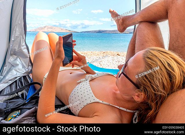 Beautiful young woman leaning on her partner while reading on her mobile phone, resting on the beach together and squeezing in the shade