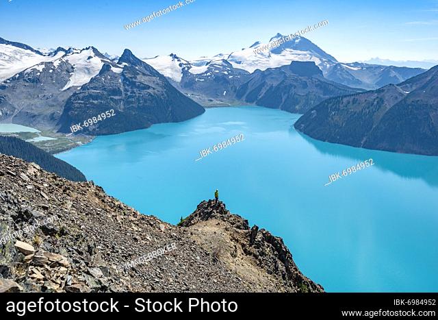Young man standing on a rock, looking into the distance, view of mountains and glacier with turquoise blue lake Garibaldi Lake, peaks Panorama Ridge