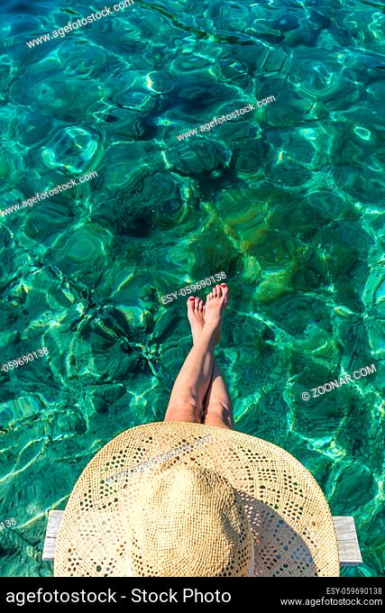 Graphic image of top down view of woman wearing big summer sun hat relaxing on small wooden pier by clear turquoise sea