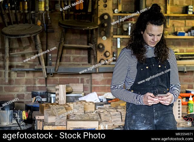 Woman with long brown hair wearing dungarees standing in wood workshop, using mobile phone