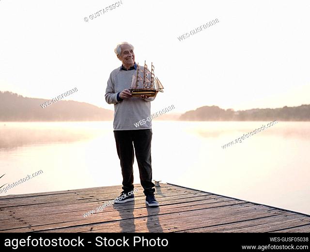Smiling senior man holding sailing ship while standing on pier