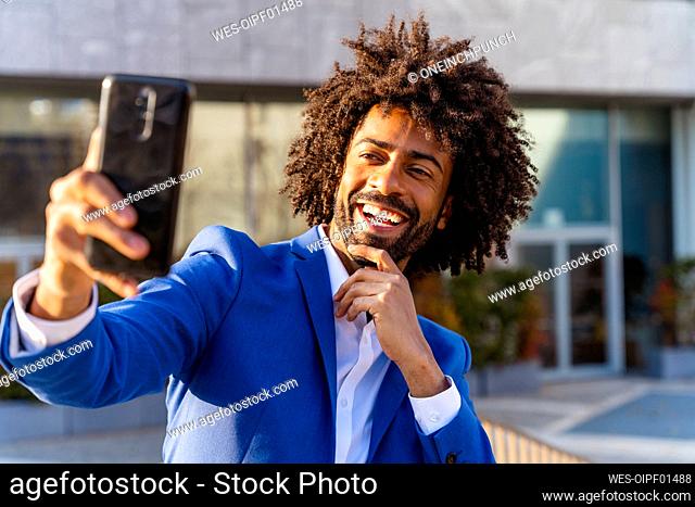 Happy businessman with hand on chin taking selfie through mobile phone in front of building