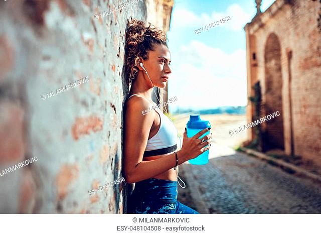 Young muscular woman with headphones standing up against the wall and resting after jogging. Looking away and thinking