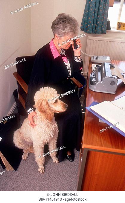 Woman with visual impairment sitting at desk in front of Braille typewriter stroking guide dog and talking on telephone