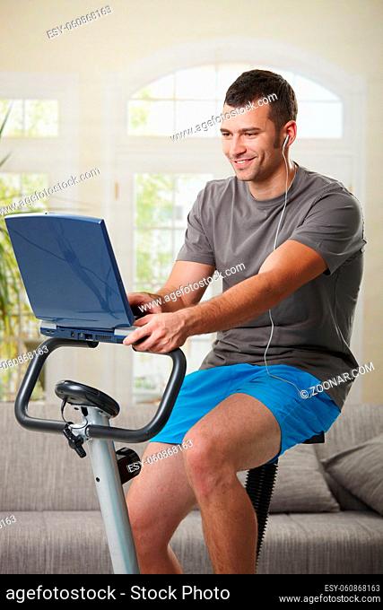 Man sitting on stationary bike at home, using laptop computer and listening music