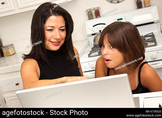 Attractive hispanic mother and mixed-race daughter in the kitchen using the laptop computer together