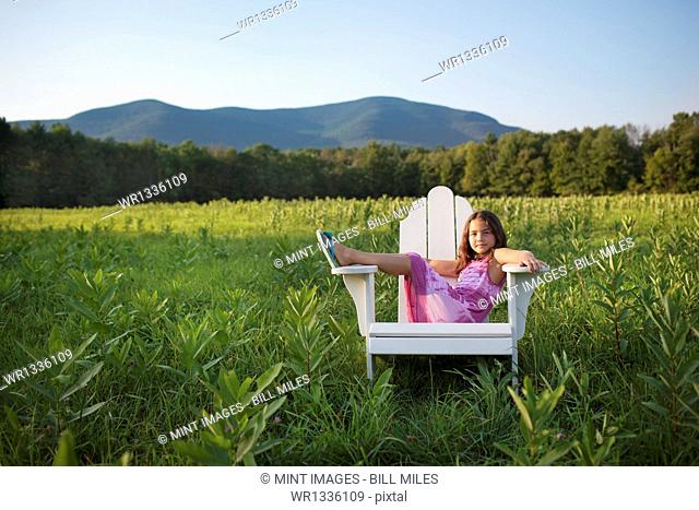 A young girl sitting in a traditional wooden Adirondack style chair in a field at evening light