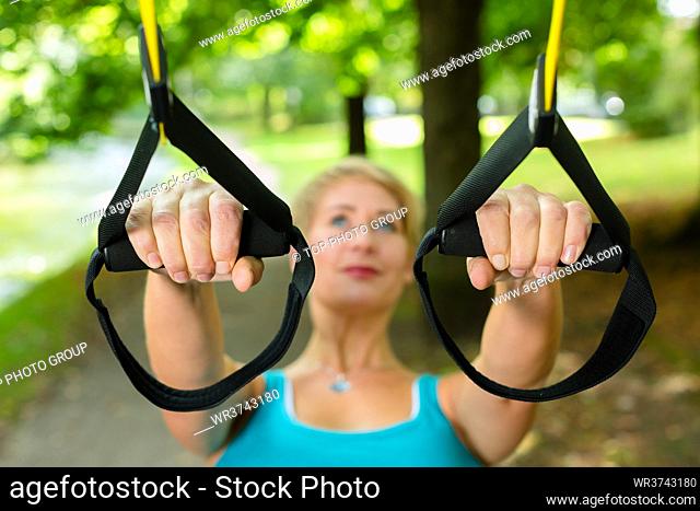Young woman exercising with suspension trainer sling in City Park under summer trees for sport fitness
