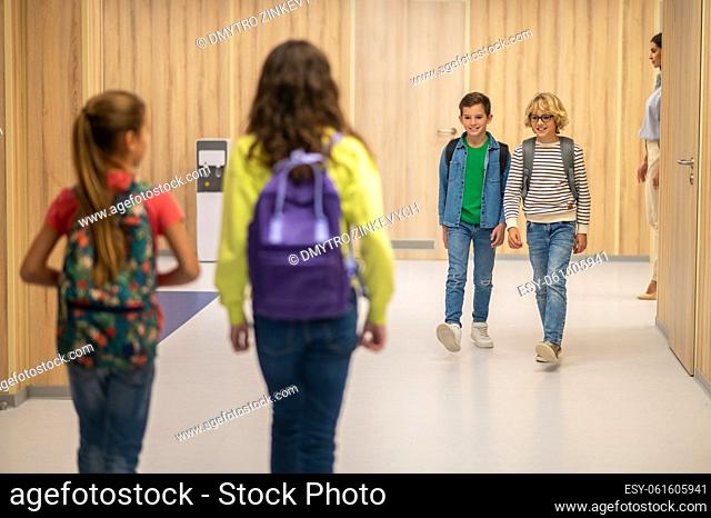 School life. Smiling boys looking at girls with backpacks with their backs to camera walking towards them in hallway of school