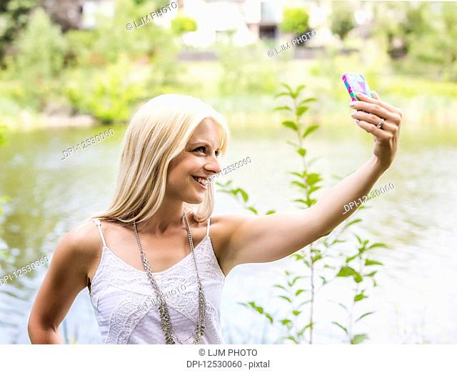 A beautiful young woman taking a self-portrait while enjoying her alone time in a park with a manmade lake; Edmonton, Alberta, Canada