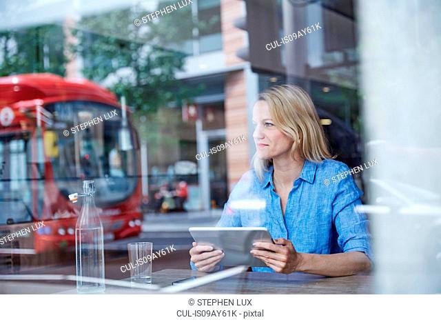 Mature woman sitting in cafe, using digital tablet, bus reflected in window