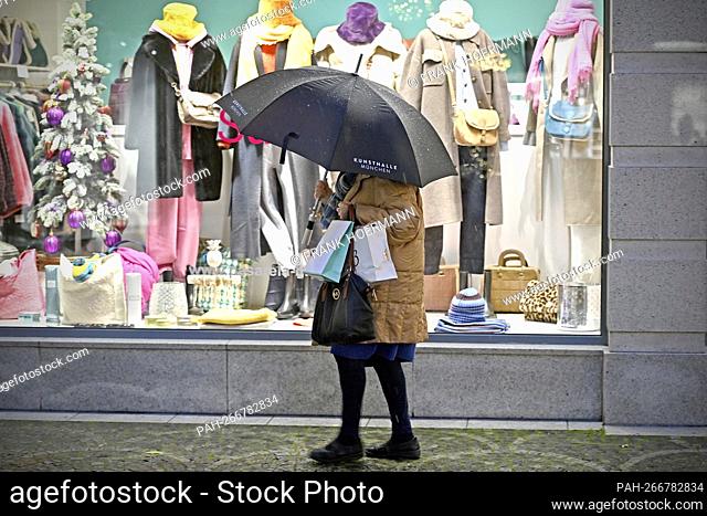 A woman stands under an umbrella with shopping bags in front of a shop window of a textile shop, fashion, clothing. - Munich/Bayern/Deutschland