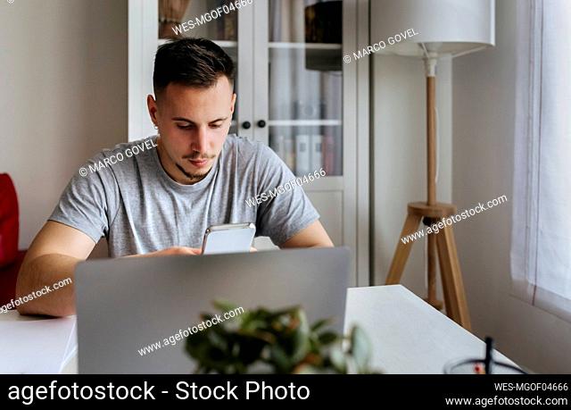 Young male entrepreneur using smart phone while sitting with laptop at home office