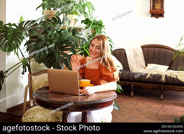 Cheerful female sitting at classic table in light living room waving hand at laptop having online call