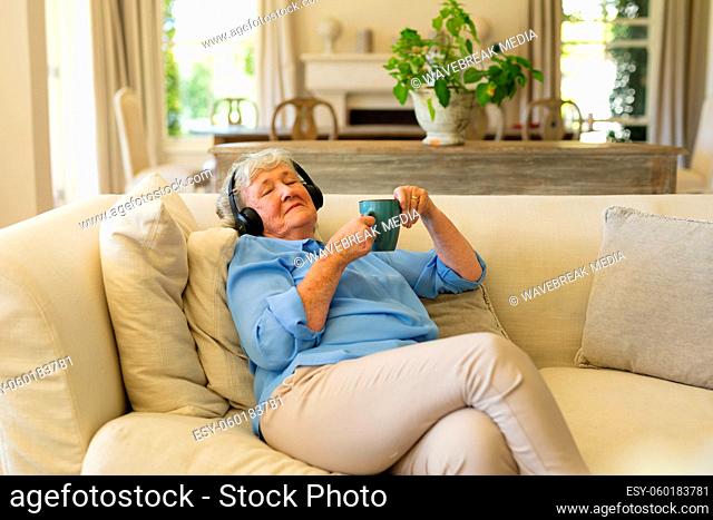 Senior caucasian woman sitting on sofa wearing headphones with eyes closed