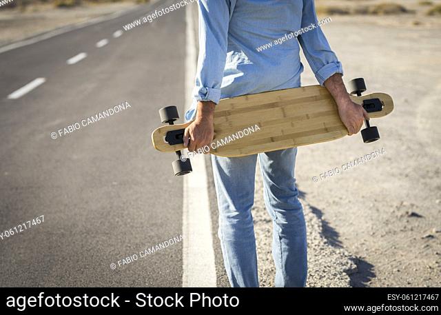 Hands of man holding skateboard on road. Rear view of man wearing denim clothes holding skateboard on rural road. Sportsman holding skateboard and waiting on...