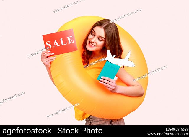 Portrait of joyful teen girl with rubber ring, holding card with sale inscription, and looking with toothy smile, enjoying summer sales for tours