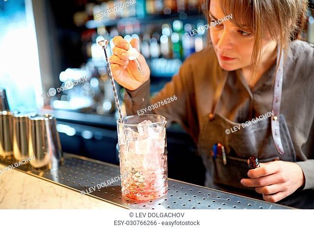 alcohol drinks, people and luxury concept - woman bartender preparing cocktail and adding essence into glass with ice at bar counter