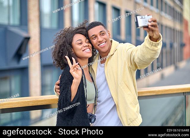 Good moments. Dark-skinned guy hugging smiling girlfriend showing freedom sign with hand taking selfie on camera on city street