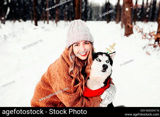 Young beautiful happy smiling woman in fur coat hugging husky dog dressed in red Christmas scarf and festive deer horns. Portrait photo of girl and dog on white...