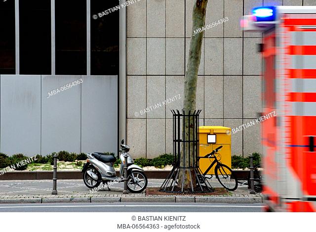 Photography of parked bicycles and a motor scooter in front of a mailbox with an ambulance with blue light driving past