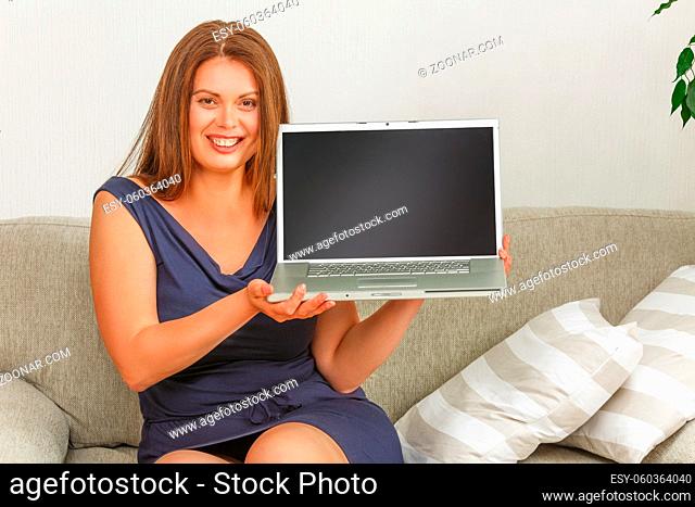Picture of beautiful businesswoman holding laptop computer in front of her and smiling for camera. Pretty lady sitting on couch at home