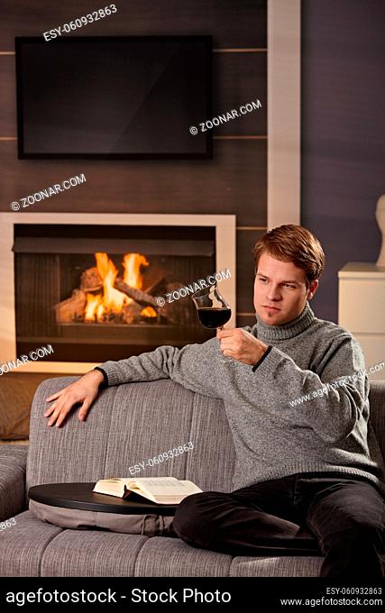 Young man sitting on sofa at home on a cold winter day, reading book in front of fireplace, tasting red wine