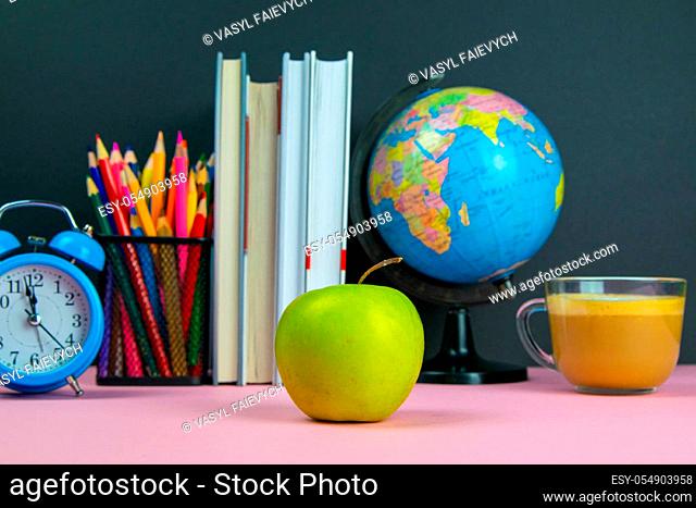 Apple is in the foreground and behind him are a stack of books, a globe, a clock and a glass of pencils