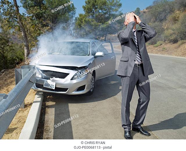 Frustrated businessman standing next to car wrecked on guardrail