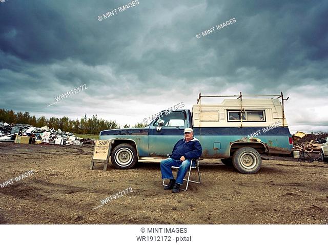 A mature man seated in a chair by his pick up truck. Piles of waste, scrap metal and wood objects